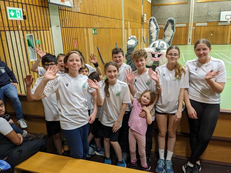 Gruppe von Jugendlichen in weißen T-Shirts, die fröhlich winken, während ein kostümiertes Kaninchen im Hintergrund steht.