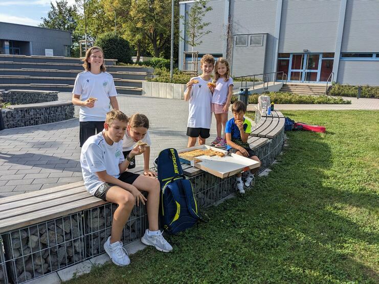 Gruppe von Kindern in Sportkleidung sitzt auf einer Bank und genießt Pizza im Freien bei schönem Wetter.