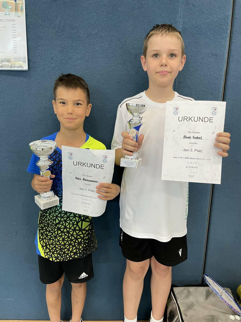 Zwei Jungen halten Trophäen und Urkunden für 1. und 3. Platz in einem Wettbewerb, vor einer blauen Wand.