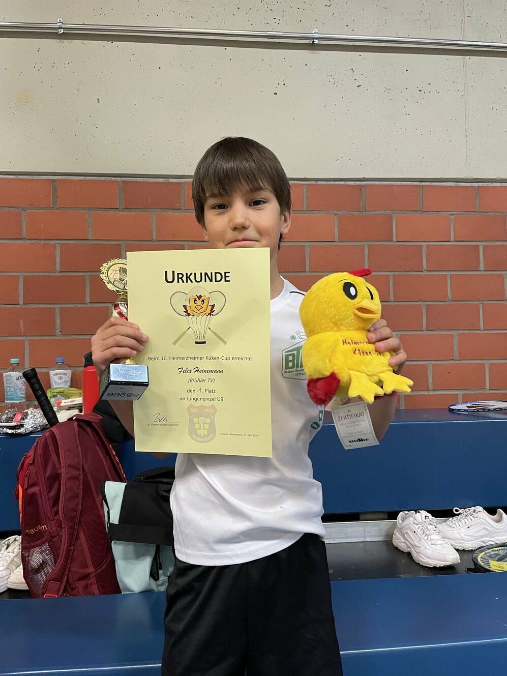 Ein Junge hält ein Diplom, einen Pokal und ein gelbes Stofftier in einer Sporthalle. Hintergrund mit Rucksack und Schuhen.