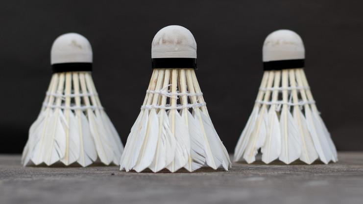 Drei Badmintonb&auml;lle aus Federn stehen in Reihe vor einem dunkelgrauen Hintergrund.