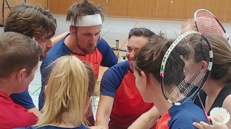 Gruppenumarmung von acht Personen im Badminton-Outfit, die motiviert vor einem Match stehen.