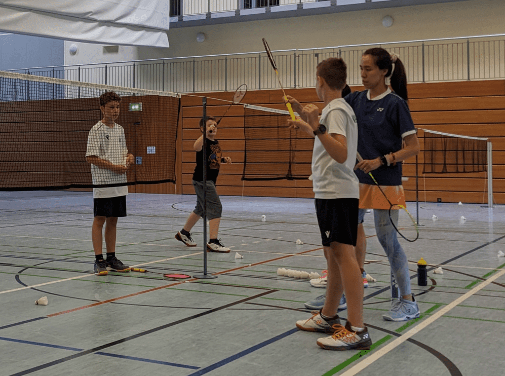 Vier Kinder mit Badmintonschlägern in einer Sporthalle, zwei besprechen etwas, während ein anderer spielt.