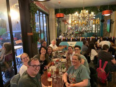 Gruppe von neun Personen in einem Restaurant, lächelnd am Tisch mit Getränken und Speisen, festliche Dekoration im Hintergrund.