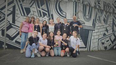 Gruppe von Mädchen und Betreuern mit Medaillen und Pokal vor einer Graffiti-Wand.