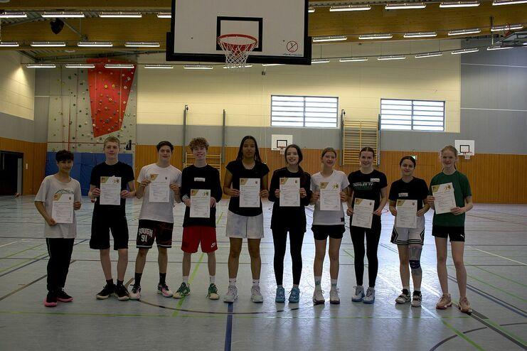 Gruppe junger Sportler hält Urkunden in einer Turnhalle mit Basketballkorb im Hintergrund.
