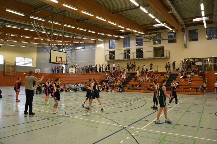 M&auml;dchenbasketballspiel in einer Halle mit Spielern in schwarz und gr&uuml;n sowie Zuschauern auf Trib&uuml;nen im Hintergrund.