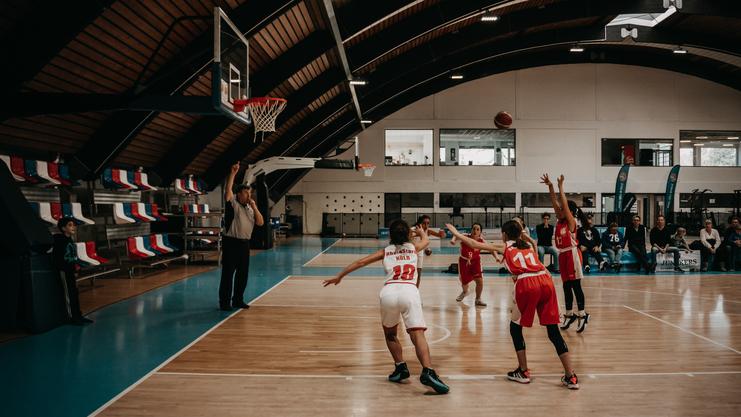 Zwei Basketballspielerinnen in roten Trikots werfen den Ball zum Korb, ein Schiedsrichter beobachtet das Spiel.