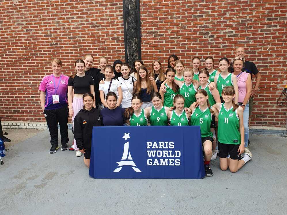 Gruppe von Mädchen in Sportkleidung, die vor einem Banner der Paris World Games posieren. Ziegelwand im Hintergrund.