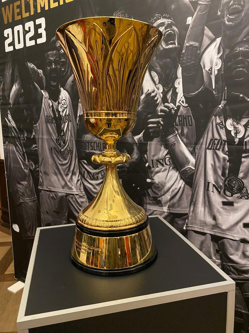 Goldener Pokal auf einem Tisch, im Hintergrund jubelnde Spieler und das Jahr 2023.