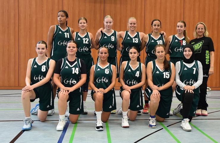 Gruppenfoto eines Basketballteams mit zw&ouml;lf Spielerinnen in gr&uuml;nen Trikots und einem Trainer auf einer Sporthalle.