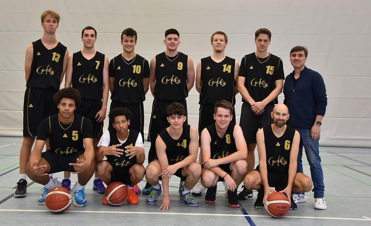 Gruppenfoto von zehn Basketballspielern in schwarzen Trikots mit Bällen, auf einem Sportfeld in einer Halle.