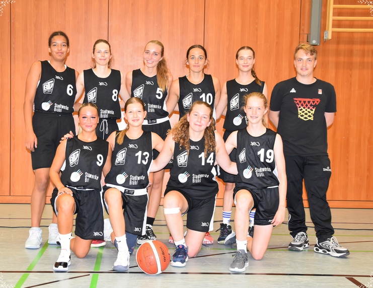 Mannschaftsfoto einer weiblichen Basketballmannschaft mit Trainer in Sportbekleidung in einer turnhallenähnlichen Umgebung.