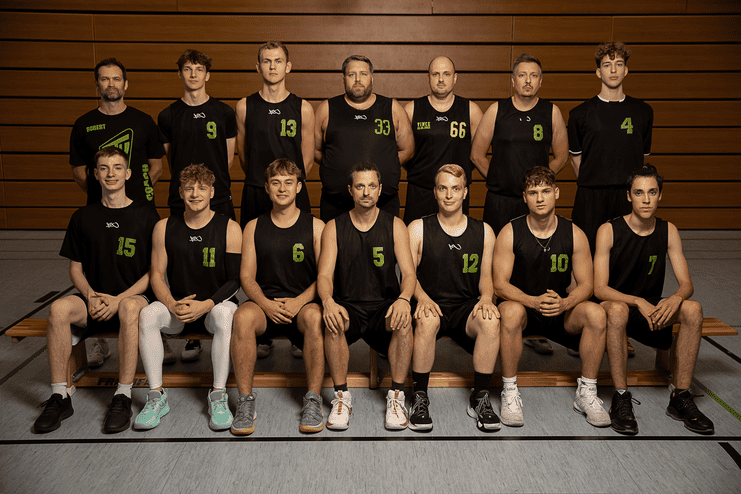 Teambild einer Basketballmannschaft mit Trainern, in Sportkleidung, auf einer Sporthalle-Bank.