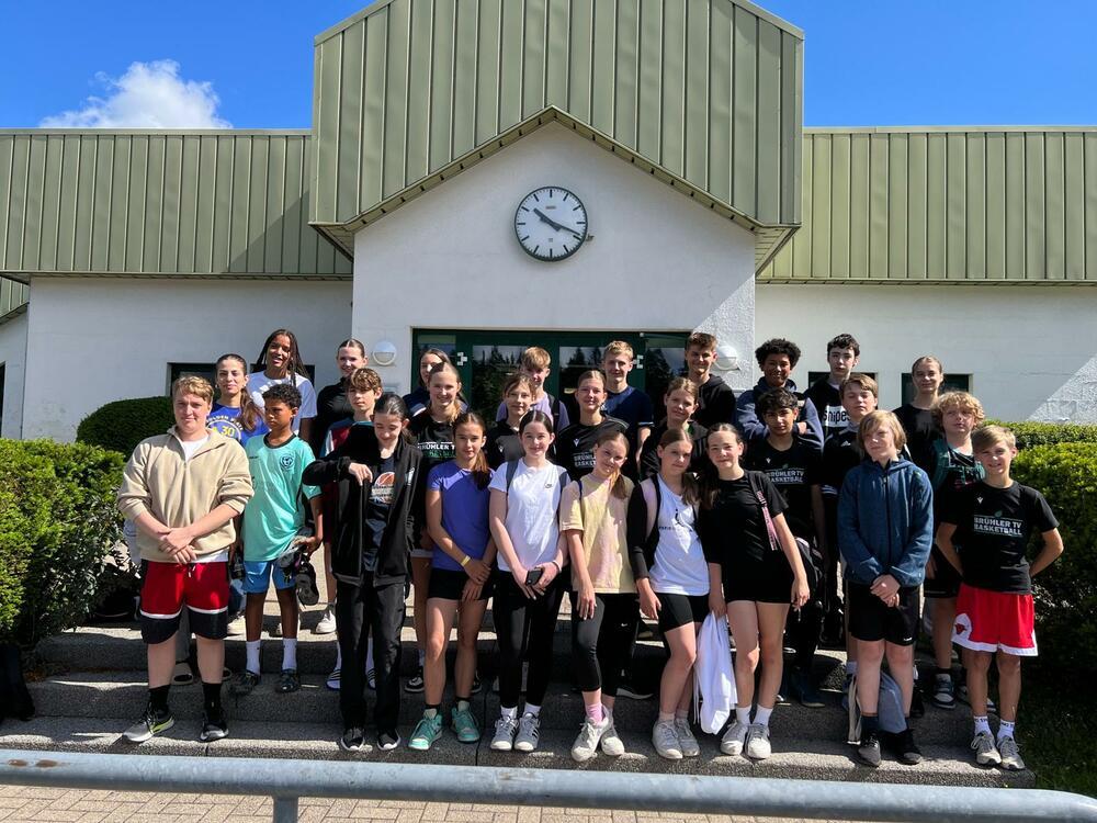 Gruppe von Jugendlichen steht vor einem Gebäude mit einem großen Uhr, umgeben von grünen Hecken und strahlend blauem Himmel.
