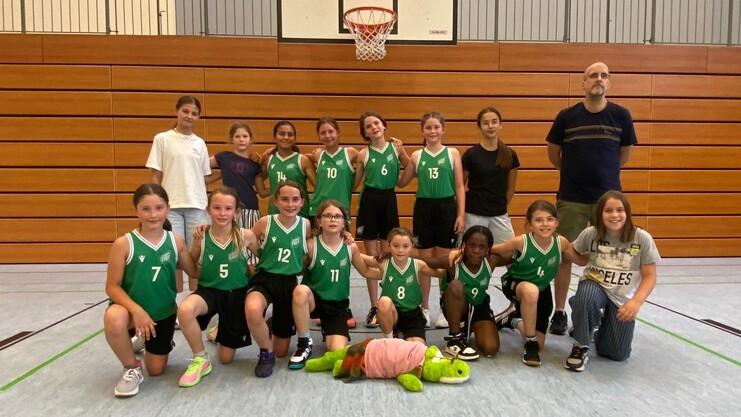 Mannschaftsphoto einer Basketballgruppe von Mädchen in grünen Trikots mit einem Trainer in einer Sporthalle.