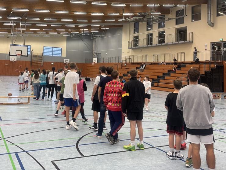 Gruppierung von Jugendlichen in Sportkleidung, die in einer Sporthalle auf eine Aktivität warten, mit Basketball und Trainingsgeräten.