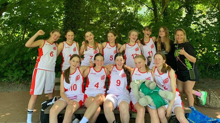 Gruppenbild von Basketballspielerinnen in roten Trikots mit einer Trainerin, umgeben von grünem Laub.