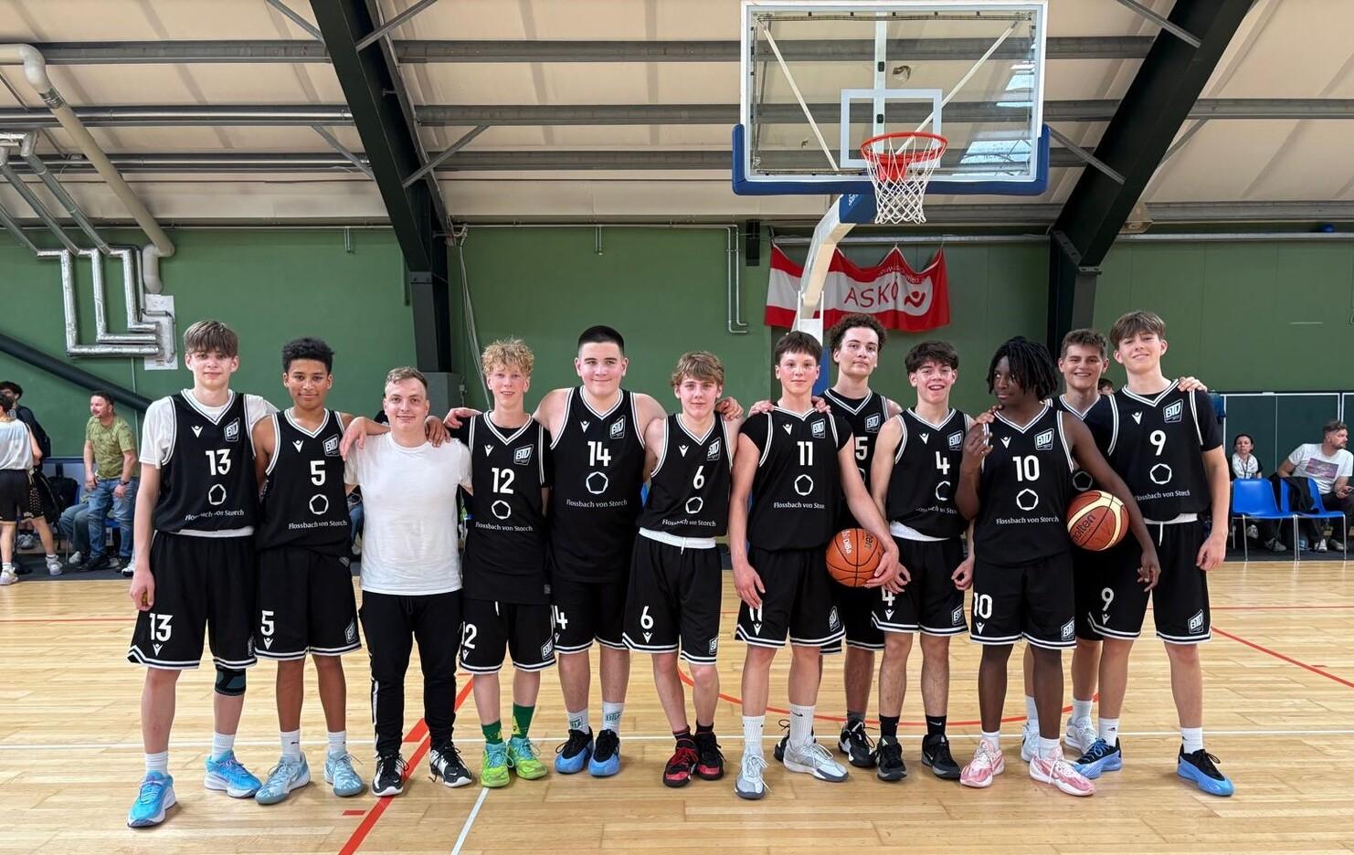 Gruppe von zwölf Basketballspielern in schwarzen Trikots, posierend auf einem Basketballfeld mit Korb im Hintergrund.