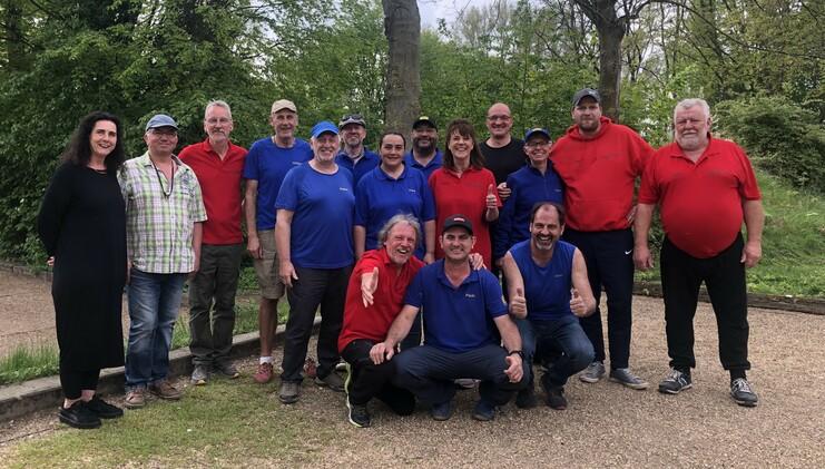 Gruppe von 15 Menschen in blauen und roten Shirts, lächelnd, im Freien vor grünen Bäumen, aufgenommen in einem Park.