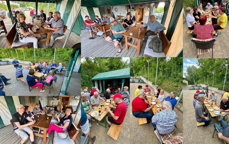 Gruppenszene mit mehreren Menschen beim Essen und Trinken unter einem Pavillon, umgeben von Bäumen und einer Sportanlage.