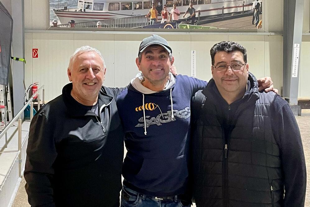 Drei Männer stehen lächelnd nebeneinander in einem modernen Innenraum mit Bootsanleger im Hintergrund.