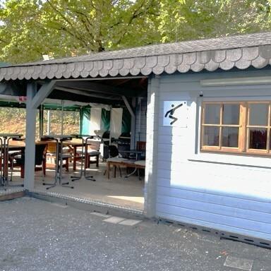 Holzhütte mit Terrasse und grüner Markise, umgeben von Bäumen, bietet Sitzmöglichkeiten im Freien.