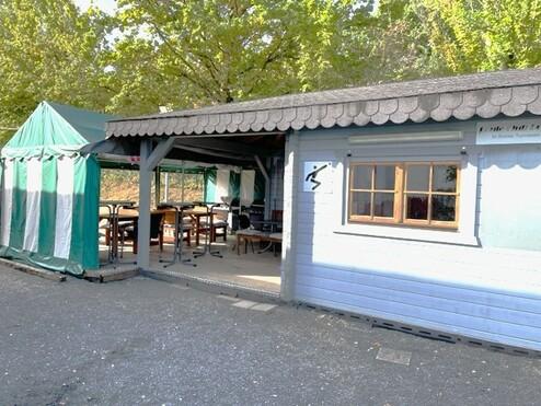 Holzhütte mit Terrasse und grüner Markise, umgeben von Bäumen, bietet Sitzmöglichkeiten im Freien.