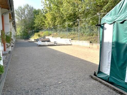 Blick auf einen Kiesplatz mit Bänken und einem Zelt, umgeben von Bäumen und einem einzäunten Bereich.