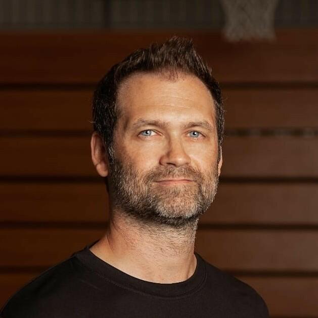 Portrait eines Mannes mit Bart in einem schwarzen Shirt mit grüner Aufschrift, im Hintergrund ein Basketballkorb.