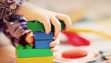 Kind baut mit bunten LEGO-Steinen einen Turm auf einem Teppich, umgeben von weiteren Spielzeugen.