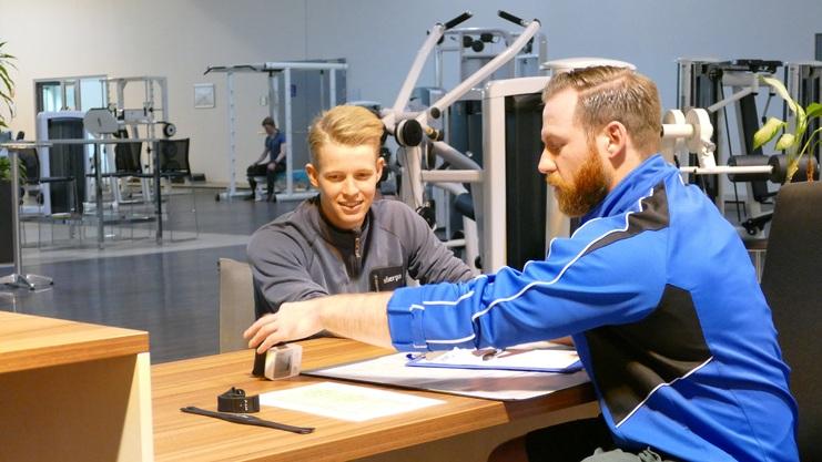 Zwei Männer am Tisch in einem Fitnessstudio, einer zeigt auf eine Uhr, während der andere interessiert schaut.
