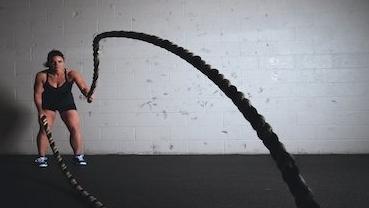 Eine Frau trainiert mit einem Battle Rope in einem Fitnessstudio vor einer grauen Wand.