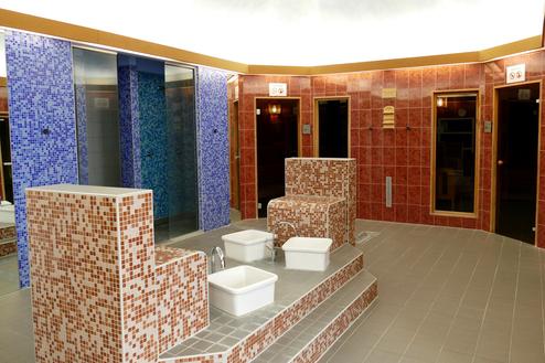 Blick ins Wellnessareal mit zwei Saunen, Duschen und Fußbecken, umgeben von bunten Fliesen auf hellen Böden.