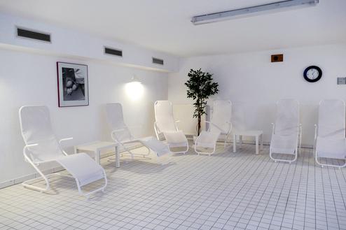 Entspanntes Ruhezimmer mit weißen Liegen, Tisch und Pflanze auf gefliestem Boden. An der Wand ist ein Bild und eine Uhr.