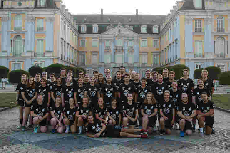 Gruppe von 50 Menschen in schwarzen T-Shirts posiert vor einem historischen, gelb-weißen Schlossgebäude.