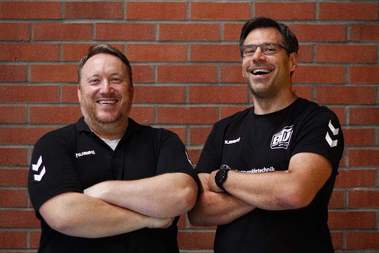 Zwei lächelnde Männer in schwarzen Sportshirts stehen mit verschränkten Armen vor einer roten Ziegelwand.