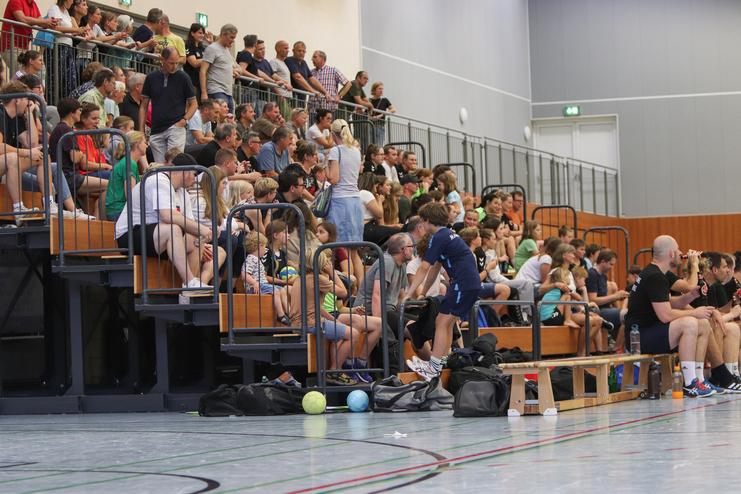 Zuschauer in einer Sporthalle sitzen auf Tribünen, während andere das Geschehen verfolgen und sich unterhalten.