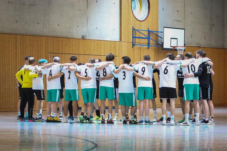 Mannschaftsversammlung im Handball, Spieler umarmen sich in einer Sporthalle vor einem Basketballkorb.