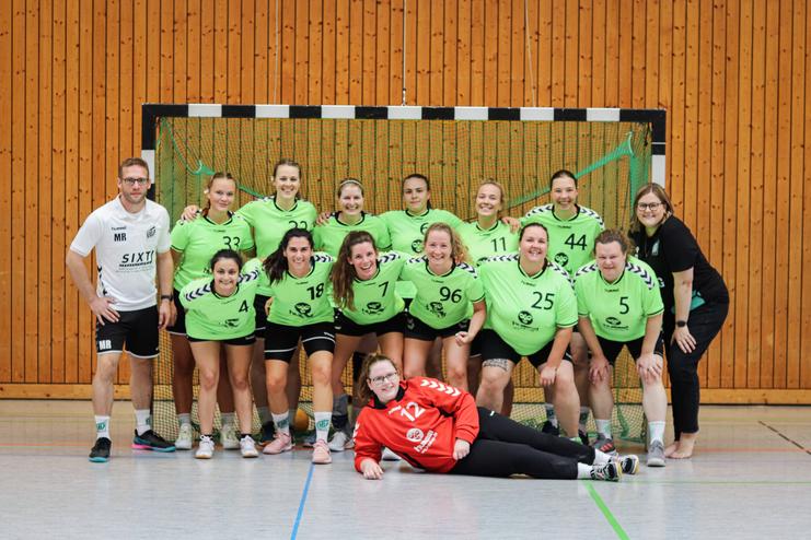 Gruppenportrait einer Frauenhandballmannschaft in grünen Trikots mit Trainer und Torhüterin auf dem Boden.