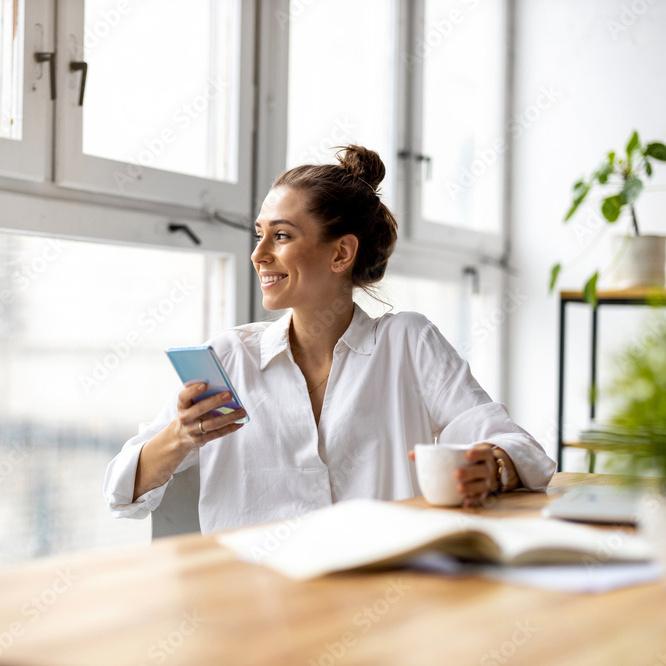 Frau mit Smartphone und Tasse sitzt am Tisch und blickt aus einem großen Fenster in einem hellichten Raum.