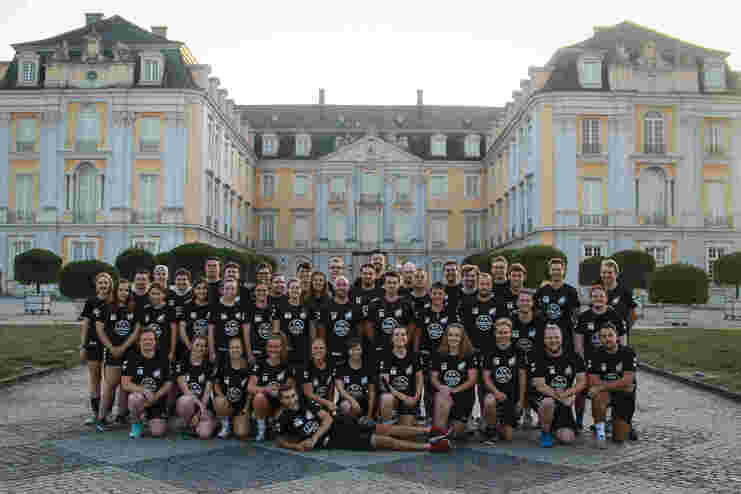 Gruppe von 50 Menschen in schwarzen T-Shirts posiert vor einem historischen, gelb-weißen Schlossgebäude.