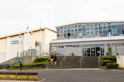 Eingang eines modernen Gebäudes mit Treppe, umgeben von Bäumen und Fahnen, an einem bewölkten Tag.