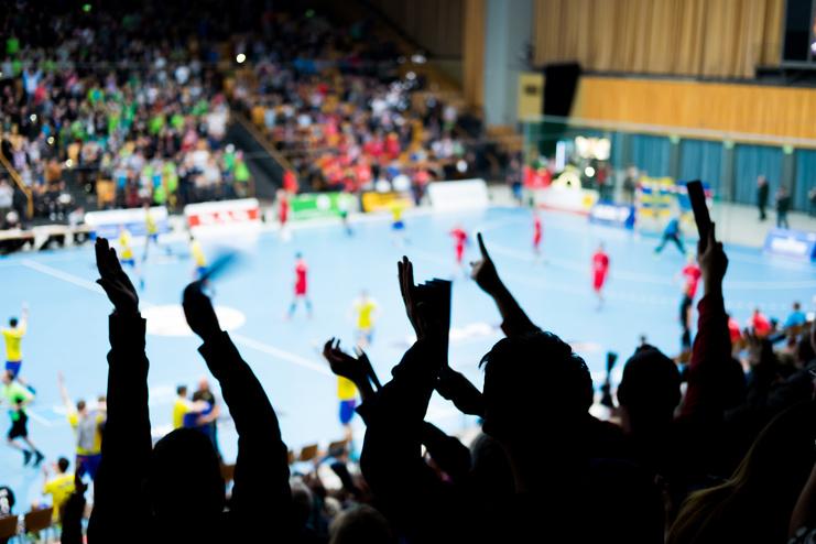 Feiernde Fans mit erhobenen Händen in einer Sporthalle während eines Handballspiels auf blauem Spielfeld.