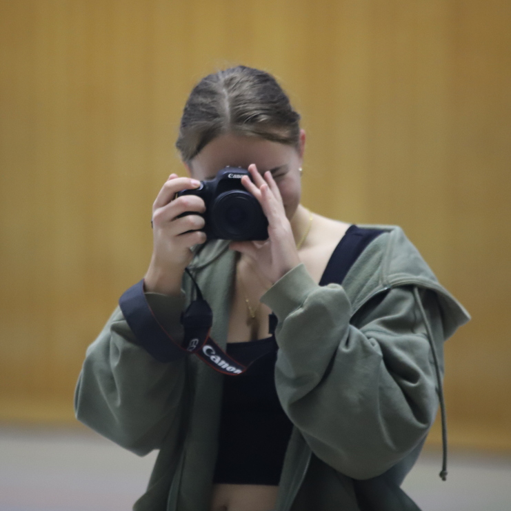 Junge Frau mit Kamera in der Hand, die in die Linse schaut, vor einer Holzvertäfelung.