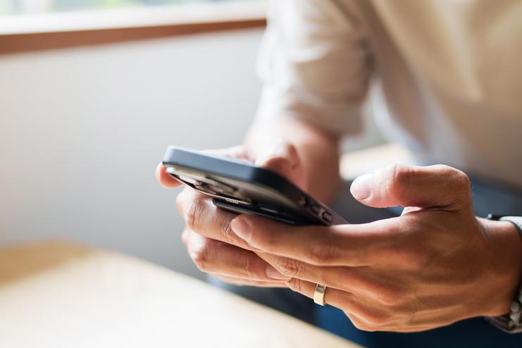 Nahaufnahme von einer Hand, die ein Smartphone hält, während sie auf den Bildschirm tippt. Holzoberfläche im Hintergrund.