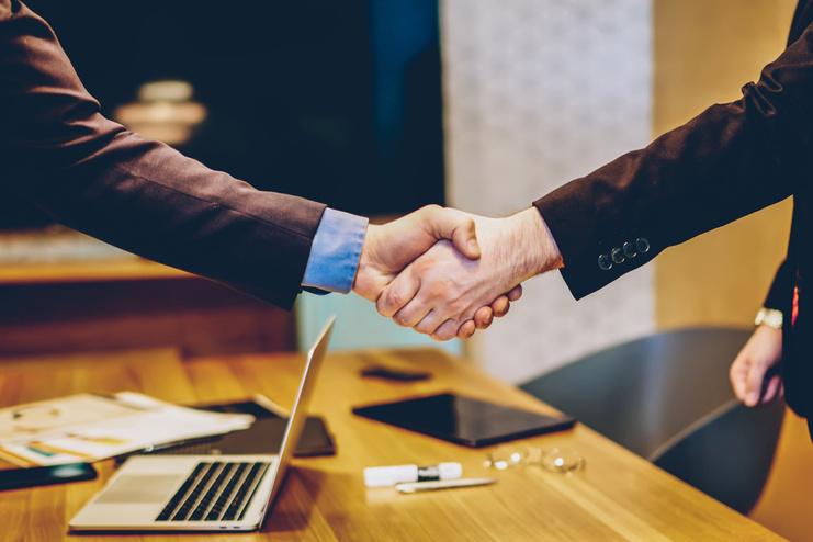 Zwei Männer in Anzügen schütteln sich die Hände über einem Tisch mit Dokumenten und einem Laptop.