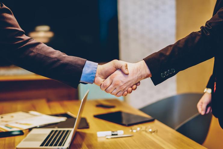 Zwei Männer in Anzügen schütteln sich die Hände über einem Tisch mit Dokumenten und einem Laptop.