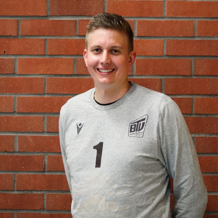 Mann im grauen Sportpulli mit der Zahl 1 steht lächelnd vor einer roten Ziegelsteinwand.