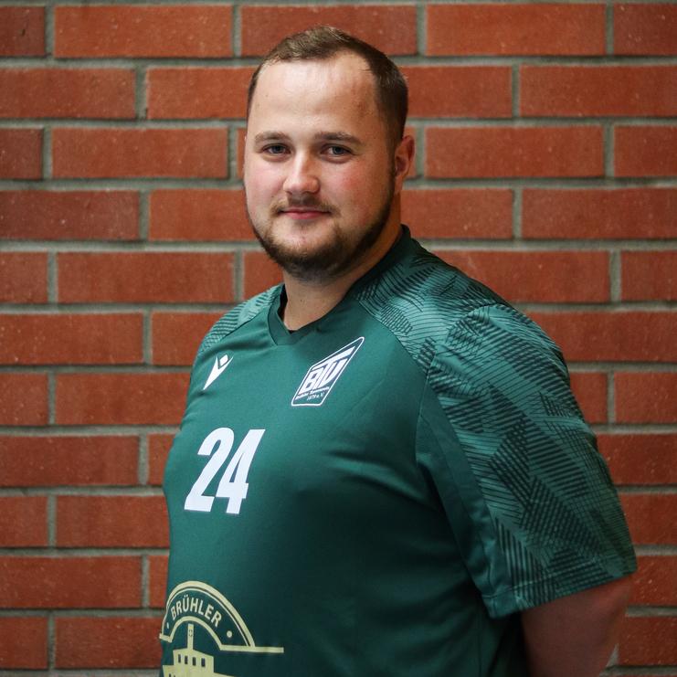 Mann in grüner Sporttrikot mit der Nummer 24 steht vor einer Ziegelsteinmauer und schaut in die Kamera.